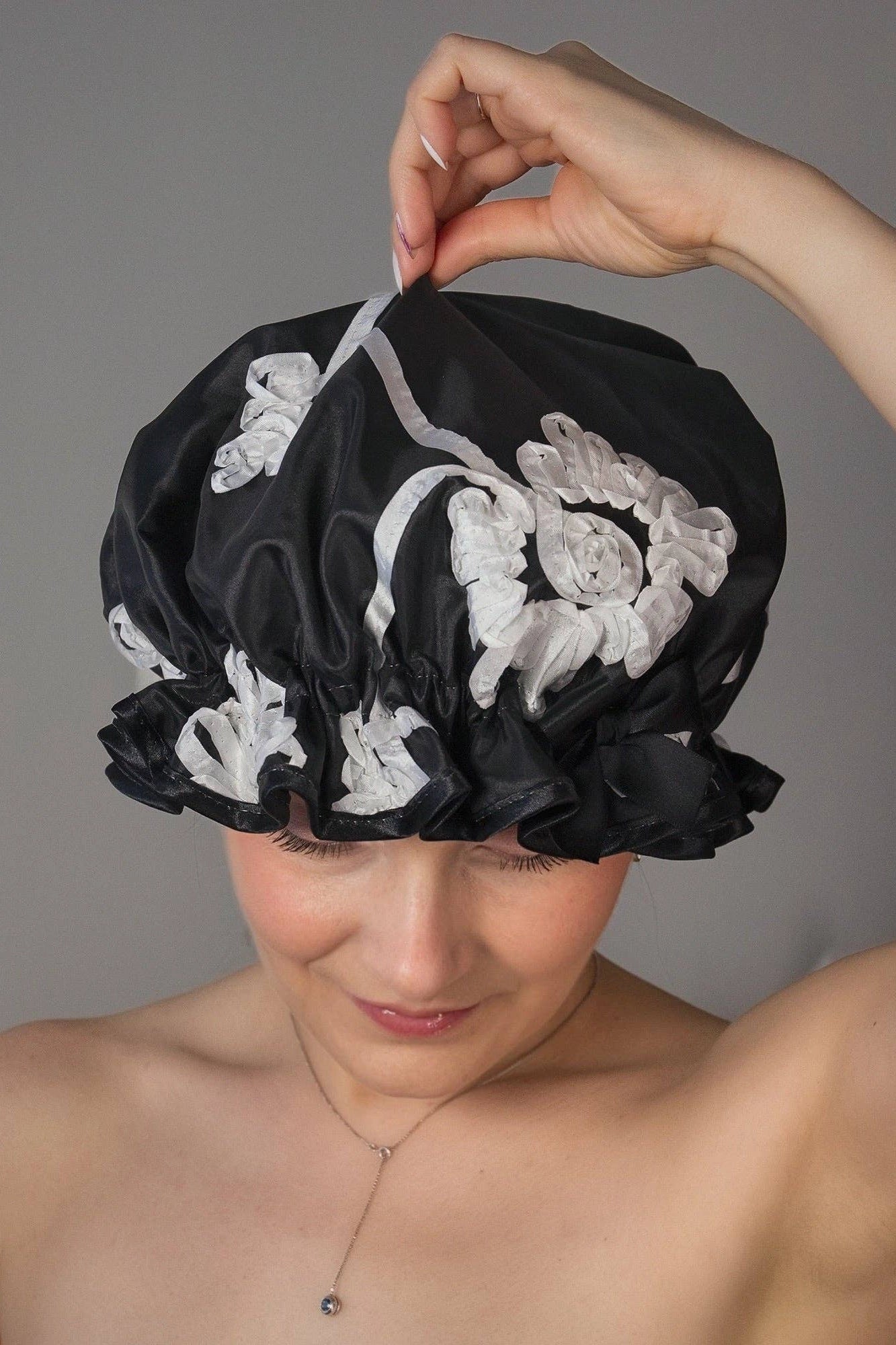 Woman wearing a lack shower cap with white ribbon floral details against a neutral background