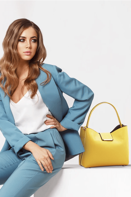 Female model with long blonde wavy hair and wearing a light blue pant suit is sitting next to the  Pebbled Leather Secchiello Bag in Pastel Yellow showing front of bag