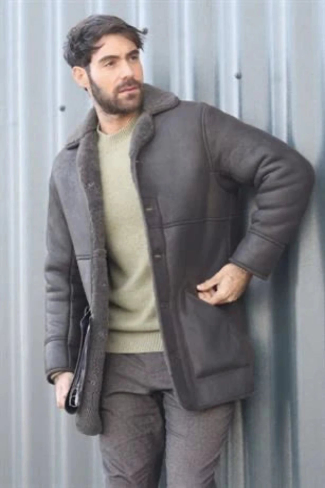 A man wearing a gray coat with two hand pockets is standing in front of a metal wall.