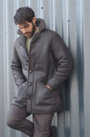 A man wearing a gray coatt with hands in  pockets is standing in front of a metal wall. looking towards ground.